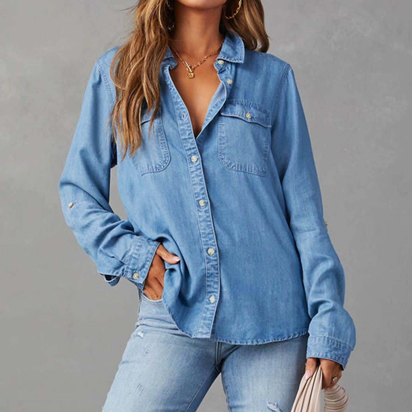 Front view of women's denim button-down shirt with collar, long sleeves, and a relaxed fit in medium blue. Shown on a model untucked, wearing distressed jeans with hand in pocket